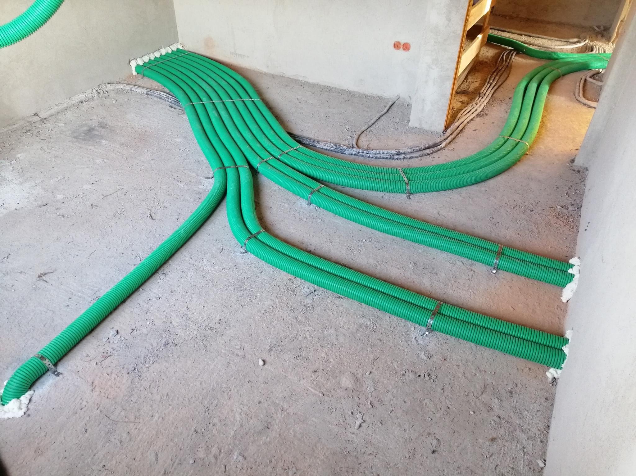 Zielone, karbowane rury wentylacyjne rozprowadzone po surowej podłodze w trakcie instalacji, zabezpieczone metalowymi opaskami i pianką montażową przy ścianach.