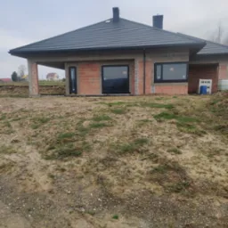 Surowy stan budynku jednorodzinnego z zamontowanymi czarnymi oknami i drzwiami zewnętrznymi, widok na ceglany mur i szary dach w pochmurny dzień.