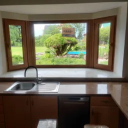 Kuchenne okno wykuszowe z drewnianą ramą, widok na zielony ogród z basenem. Wnętrze kuchni z blatem z brązowego marmuru, zlewozmywakiem i drabinką.