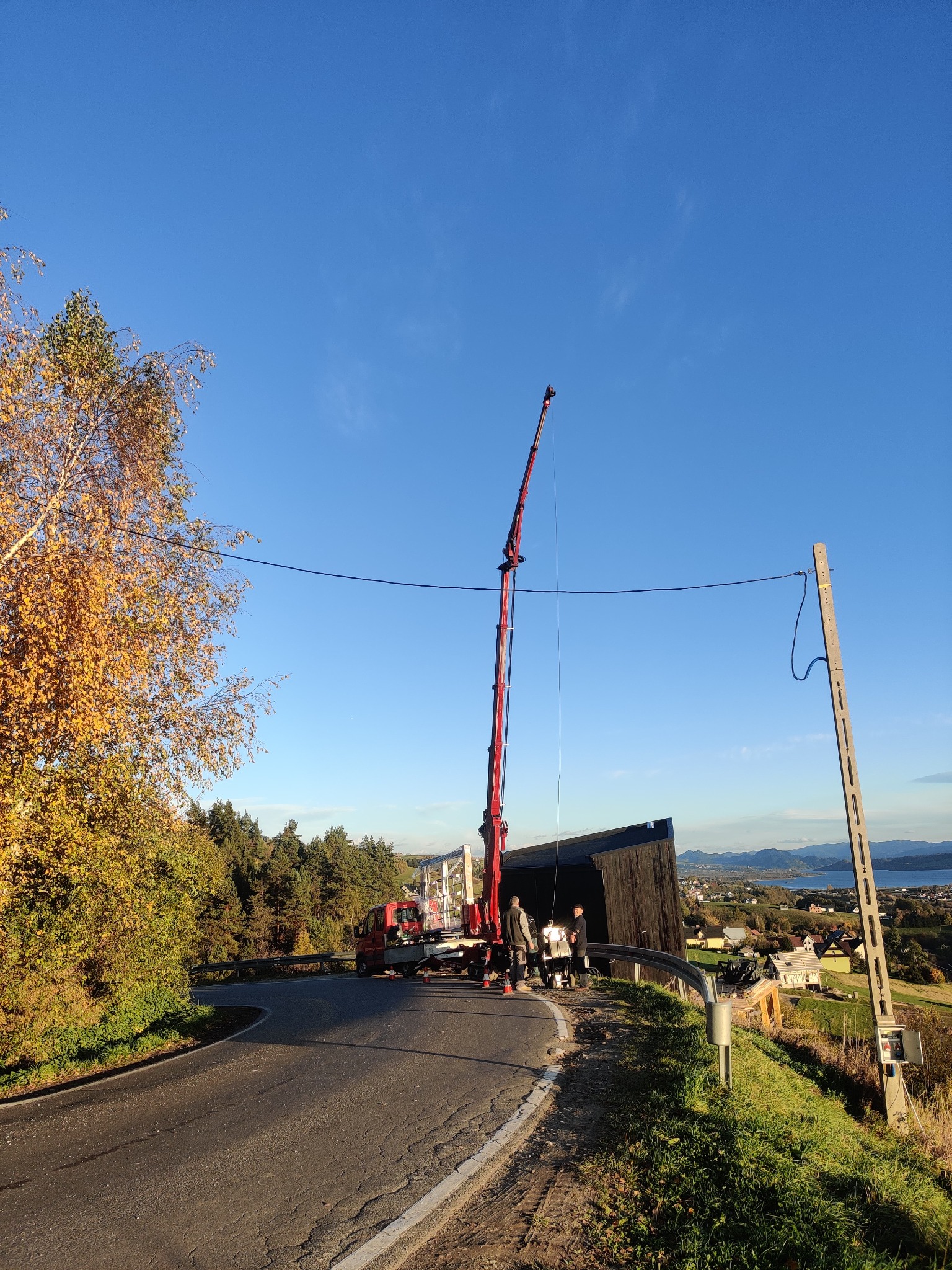 Montaż dużej, nowoczesnej, czarnej bramy garażowej z użyciem dźwigu na zakręcie drogi, w tle jesienny krajobraz i jezioro.