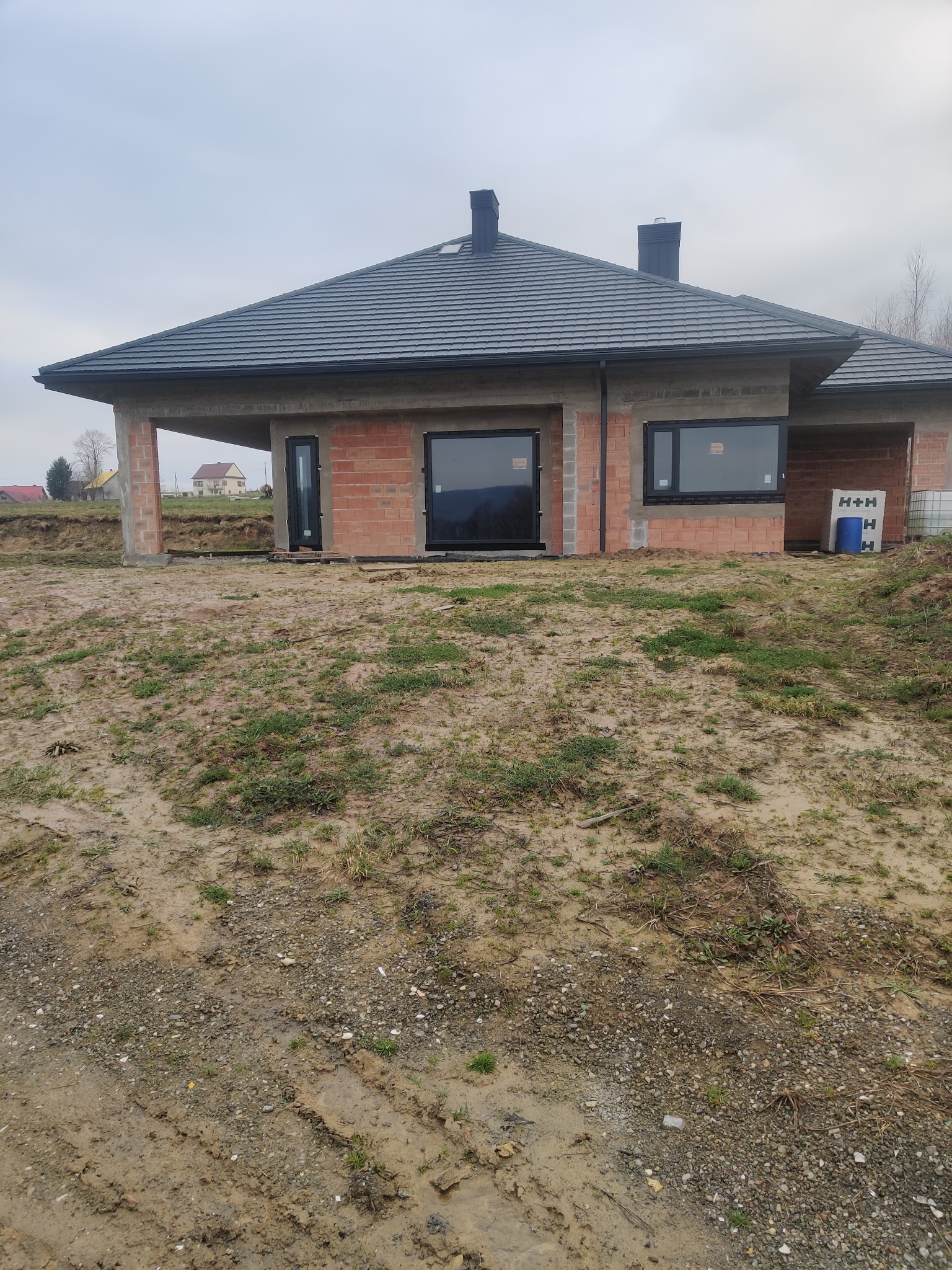 Surowy stan budynku jednorodzinnego z zamontowanymi czarnymi oknami i drzwiami zewnętrznymi, widok na ceglany mur i szary dach w pochmurny dzień.