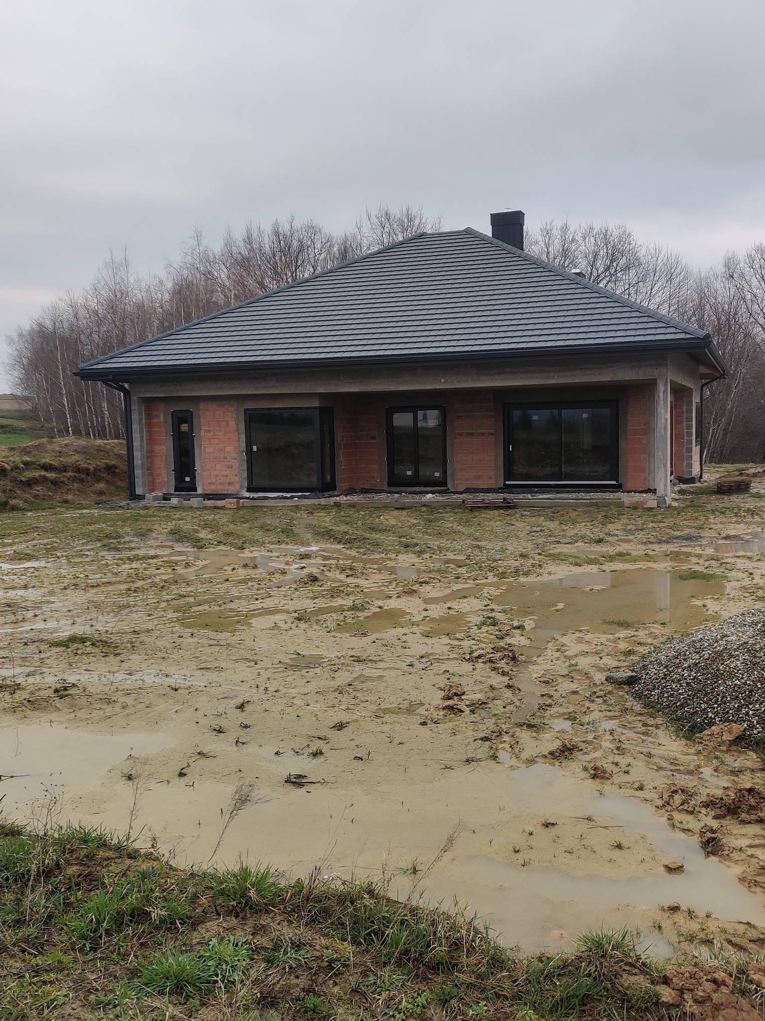 Surowy stan domu jednorodzinnego z czarnymi oknami, widoczne cegły i szara dachówka, teren wokół domu błotnisty po deszczu, pochmurne niebo.