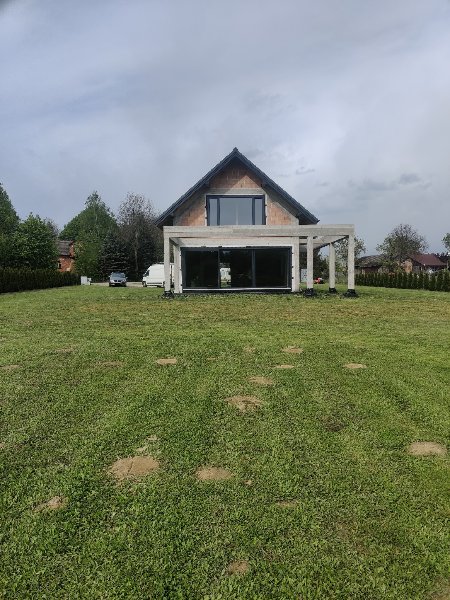 Dom w stanie surowym otwartym z dużymi, przesuwnymi oknami tarasowymi i oknem na poddaszu, widoczne samochody na posesji, duży trawnik.