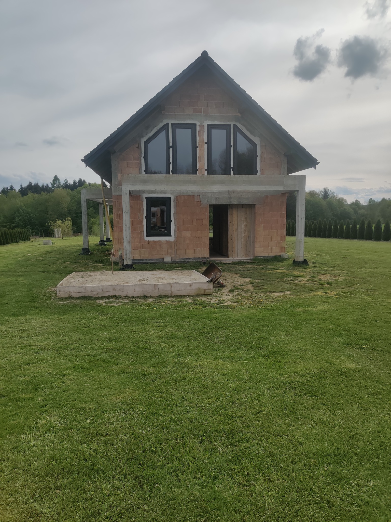 Surowy stan budynku jednorodzinnego z czarnymi oknami i otwartymi drzwiami, widoczne bloczki betonowe i zbrojenia, trawnik przed budynkiem, pochmurne niebo.