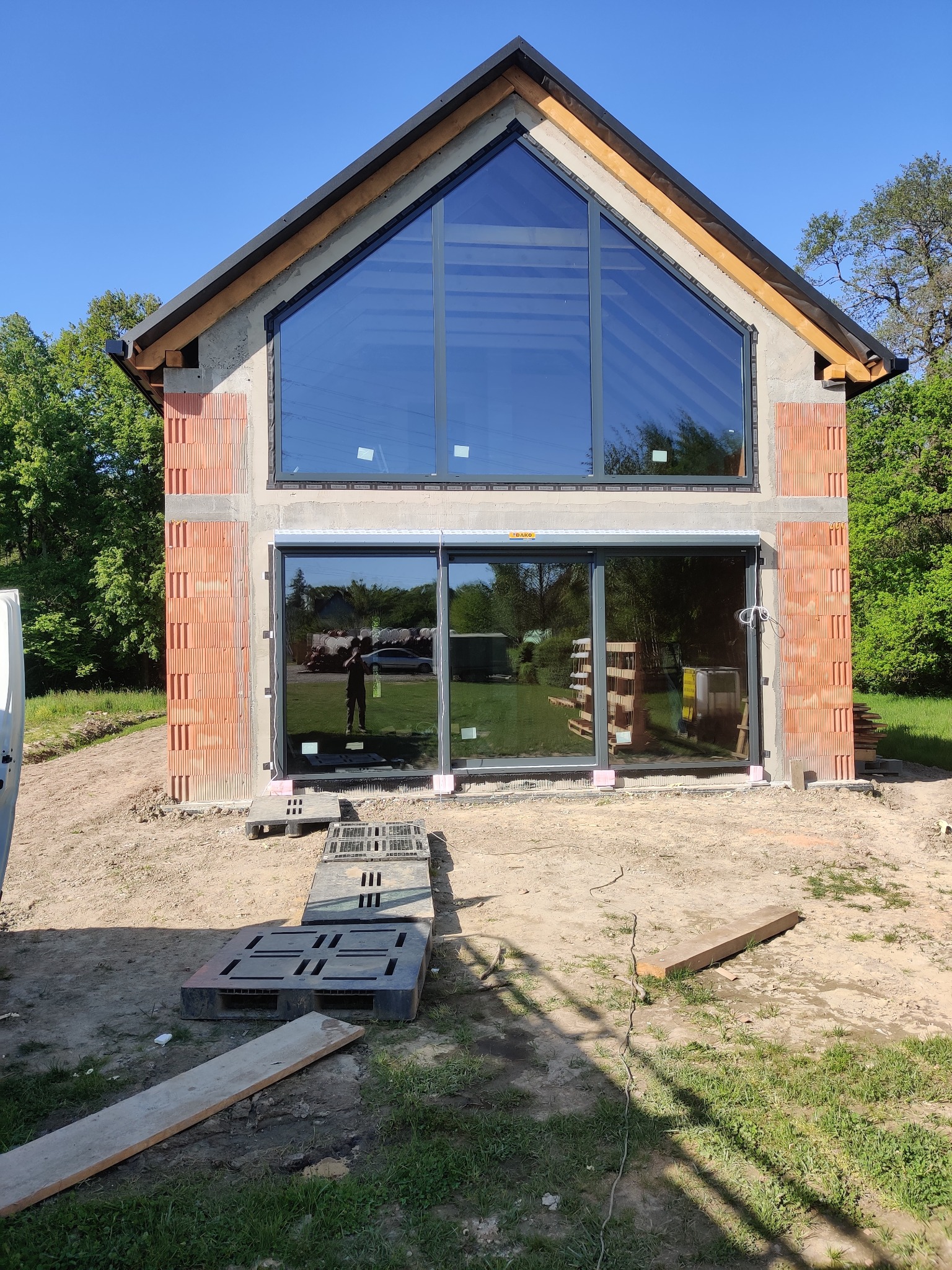 Surowy stan budynku z dużymi przeszkleniami: trójkątne okno na poddaszu i przesuwne drzwi tarasowe z szarymi ramami, widoczne cegły i palety budowlane na zewnątrz.