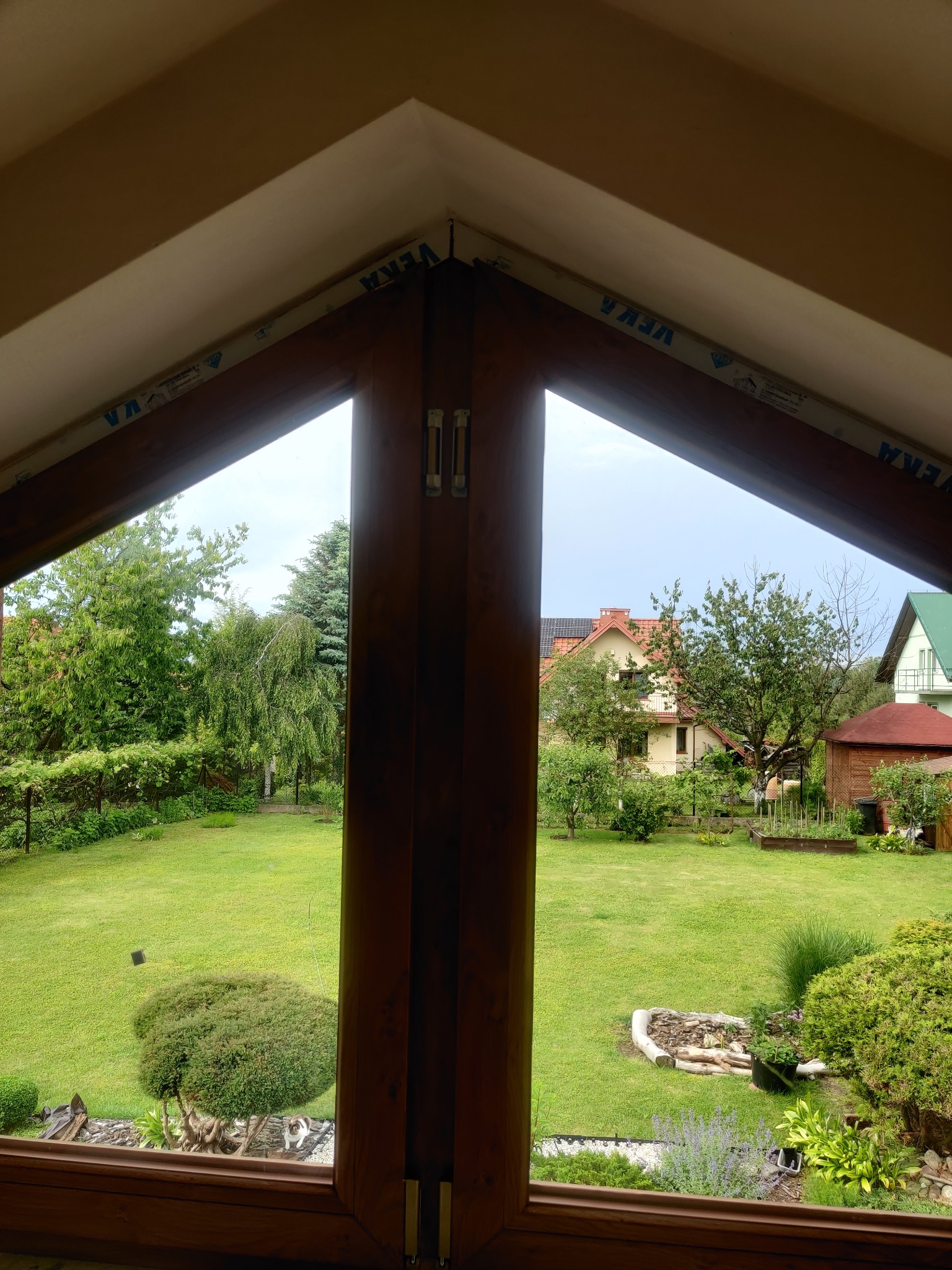 Widok z okna o nietypowym, trójkątnym kształcie na zadbany ogród z domami w tle, rama okienna z widocznym profilem marki VEKA.