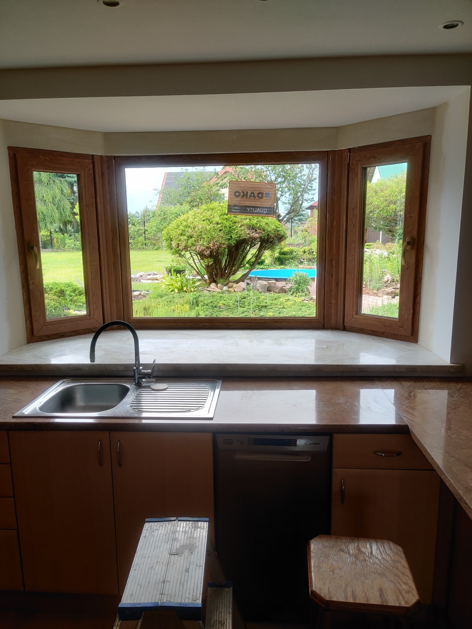 Kuchenne okno wykuszowe z drewnianą ramą, widok na zielony ogród z basenem. Wnętrze kuchni z blatem z brązowego marmuru, zlewozmywakiem i drabinką.