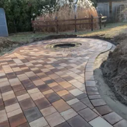 Okrągły plac z kostki brukowej w odcieniach brązu i beżu, z centralnym otworem na ognisko, otoczony ziemią i przygotowany pod trawnik.