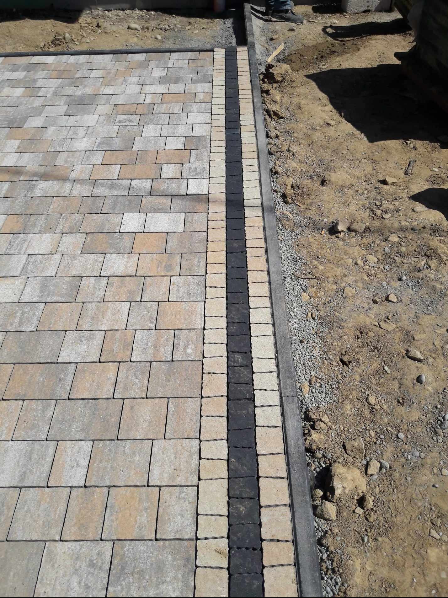 Ułożona kostka brukowa w odcieniach szarości i beżu, z ciemnym obramowaniem, na tle ziemi i gruzu.