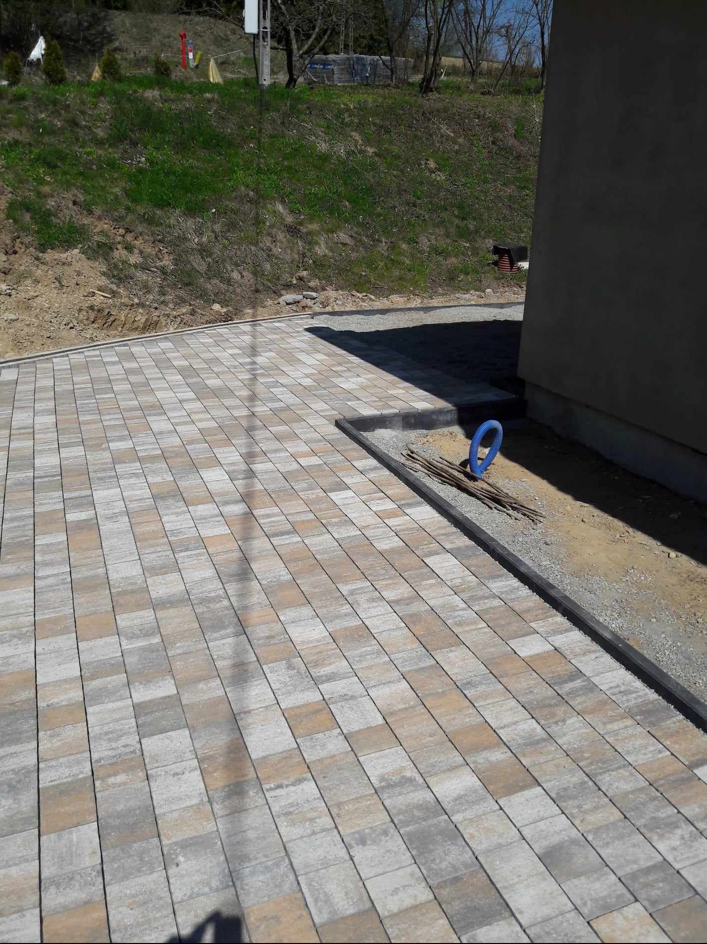 Świeżo ułożona, wielobarwna kostka brukowa na podjeździe, z widocznymi rurami i prętami zbrojeniowymi przy krawędzi, na tle zielonego wzgórza i szarej ściany budynku.