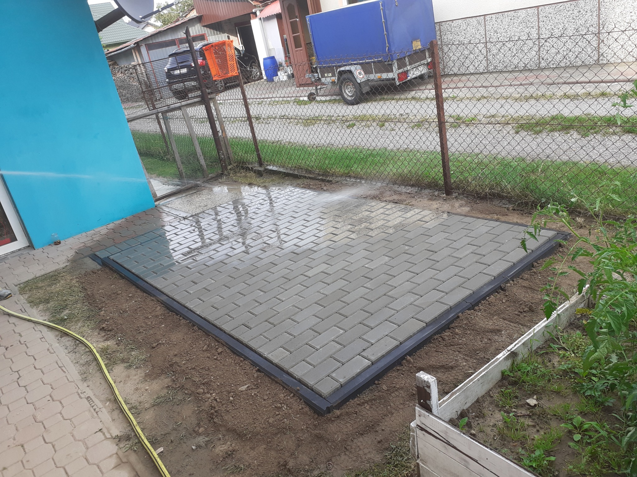 Umyta kostka brukowa ułożona na przygotowanym podłożu, obramowana czarnym krawężnikiem, widoczne ślady wody i mokra ziemia.