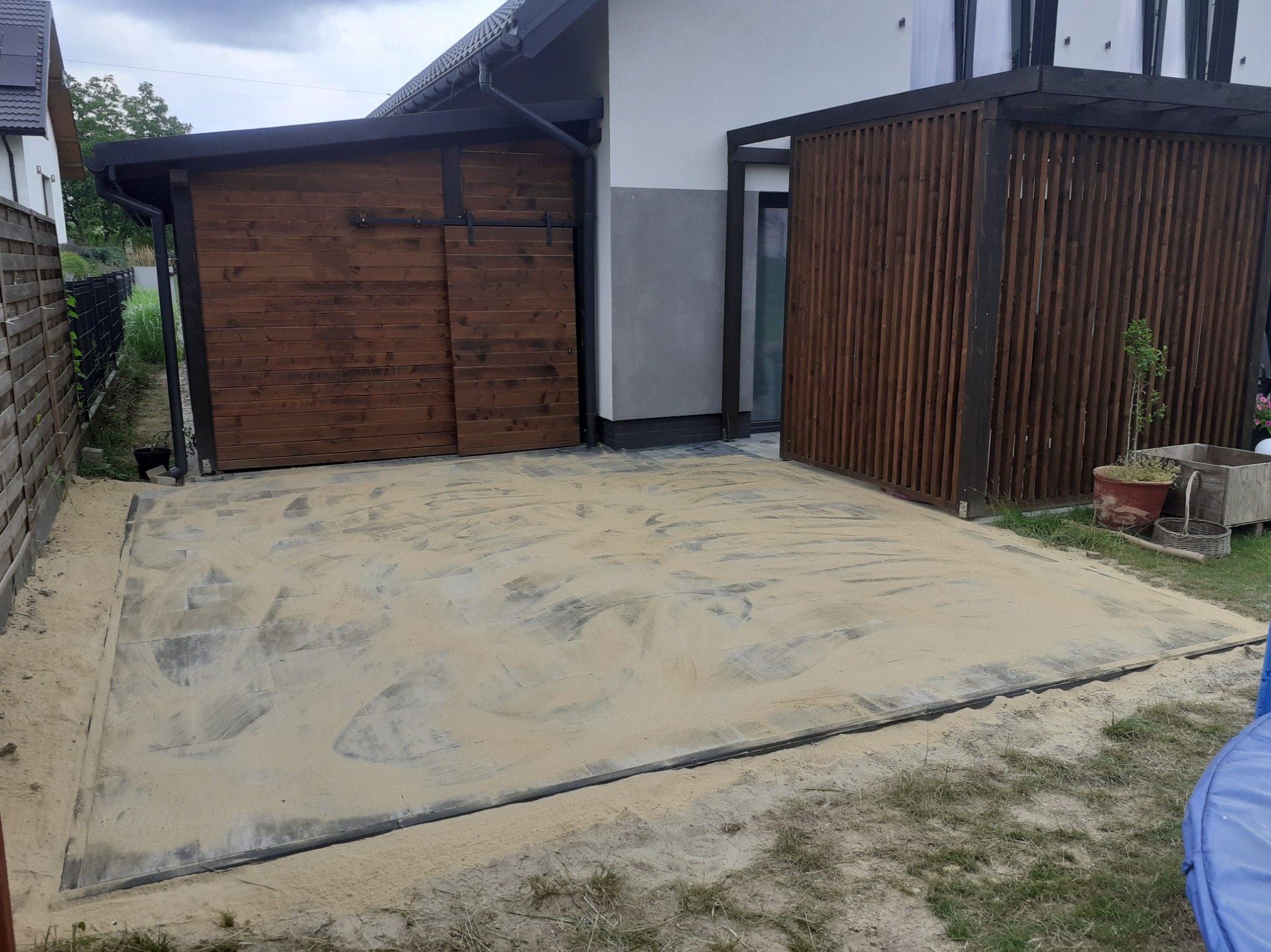Przygotowany teren pod przyszłą nawierzchnię z kostki, podsypany piaskiem, z widoczną obudową garażu z przesuwnymi drzwiami i drewnianą konstrukcją w tle.