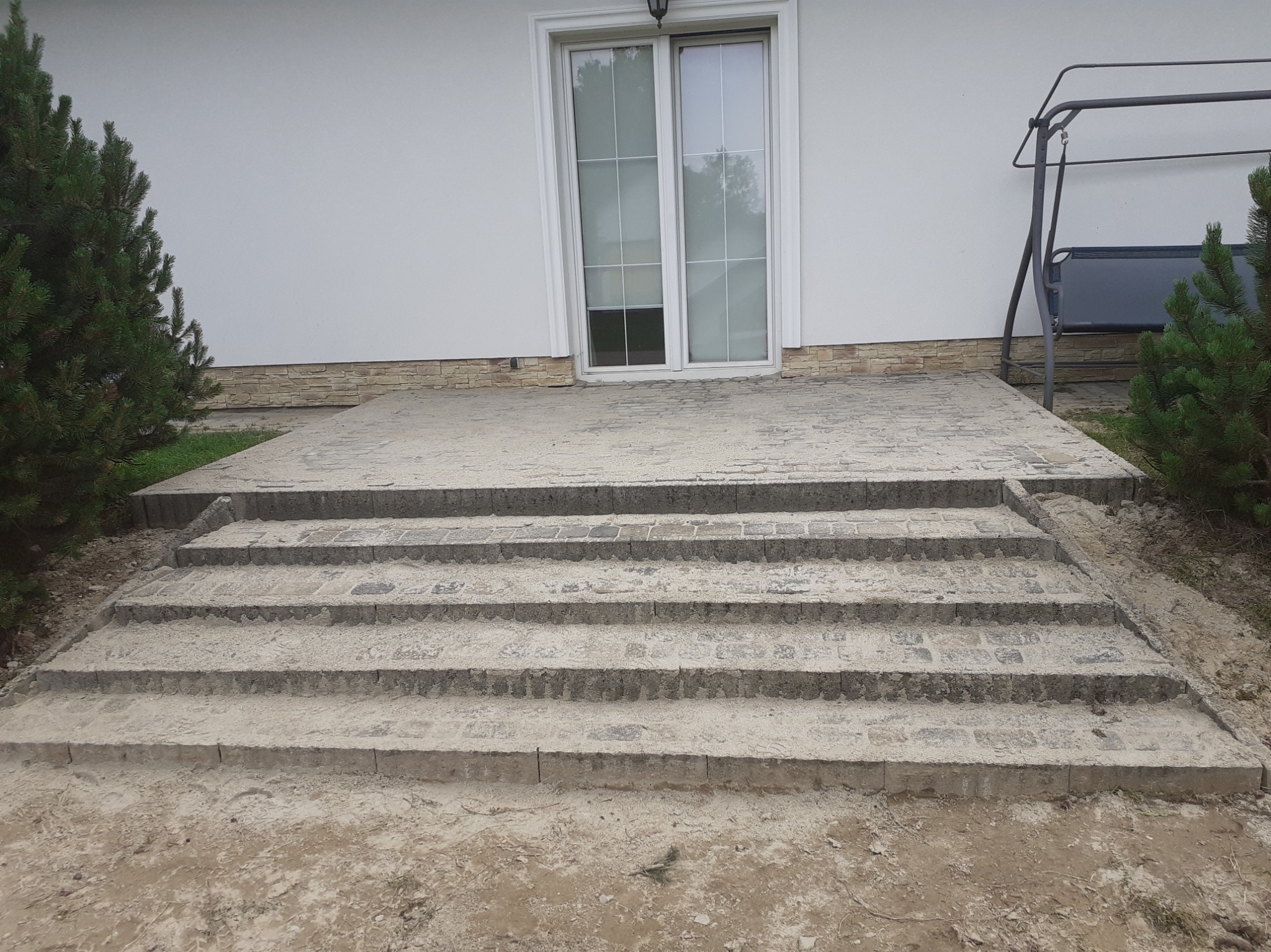 Schody wejściowe do domu wyłożone szarą kostką brukową, z widocznym fragmentem elewacji i roślinnością.