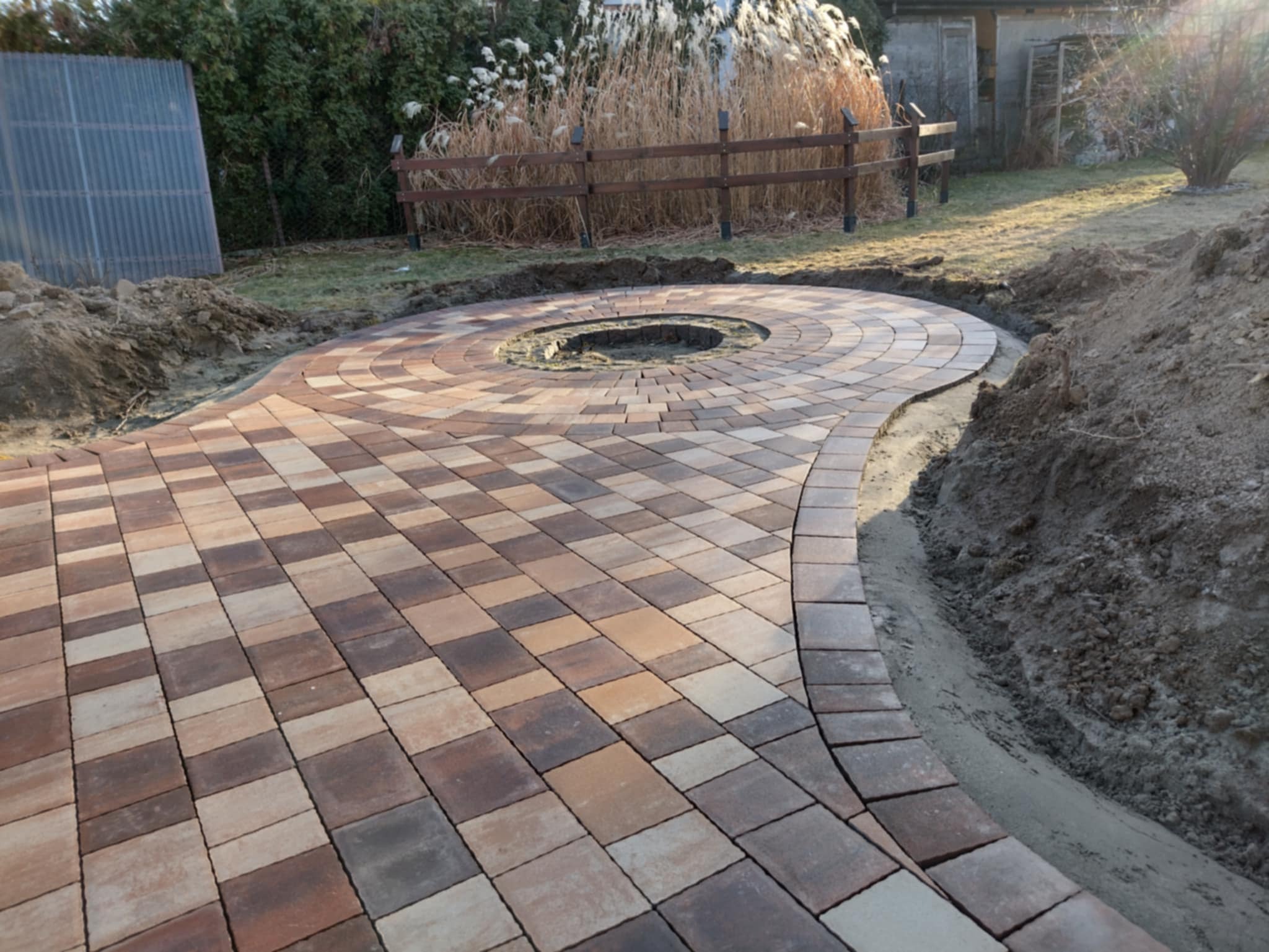 Okrągły plac z kostki brukowej w odcieniach brązu i beżu, z centralnym otworem na ognisko, otoczony ziemią i przygotowany pod trawnik.