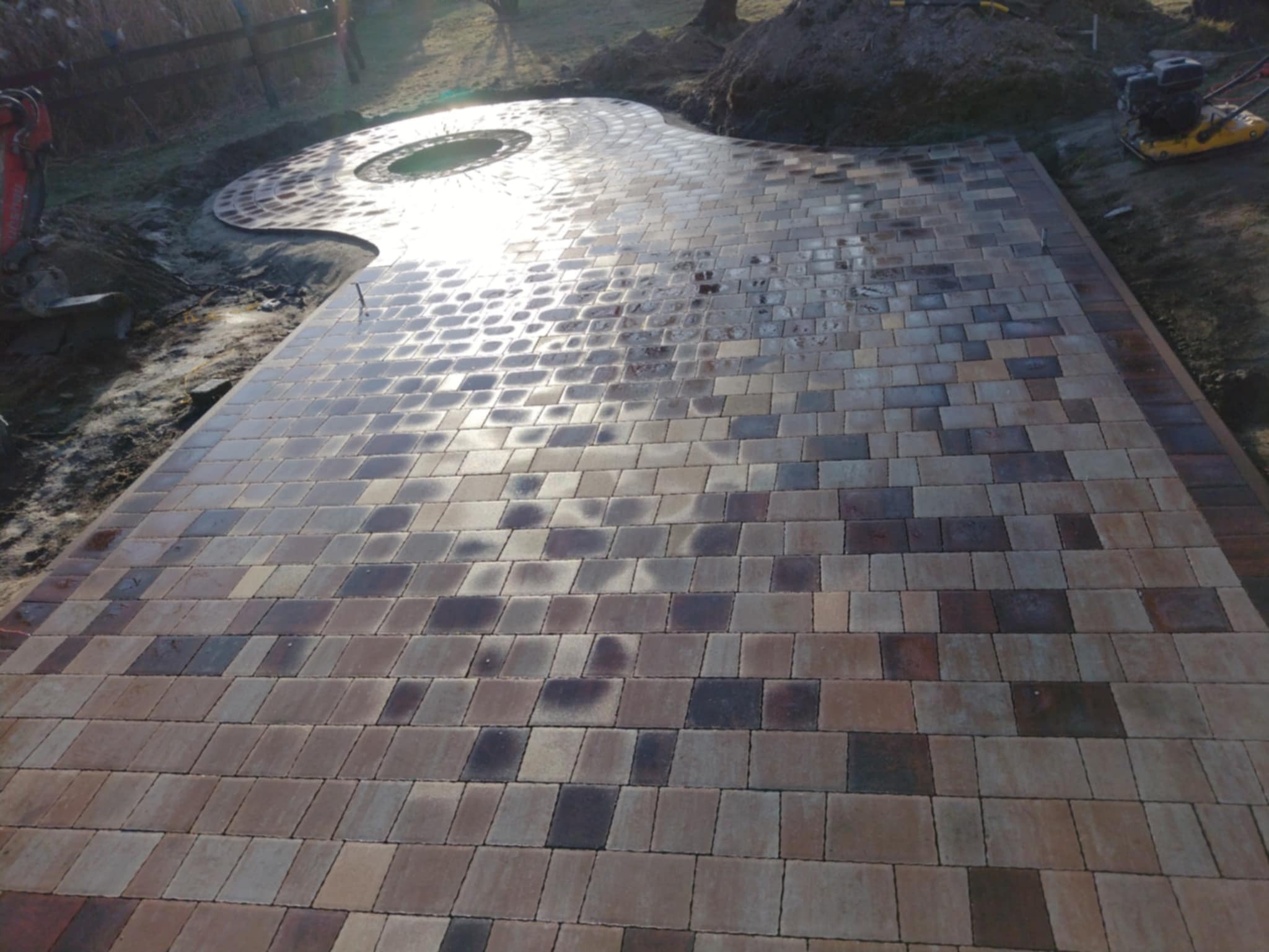 Nowo ułożona, wielobarwna kostka brukowa w odcieniach brązu i szarości, układająca się w prostokątną ścieżkę i okrągły element z otworem pośrodku, widoczna ubijarka na ziemi i rozkopany teren w tle.