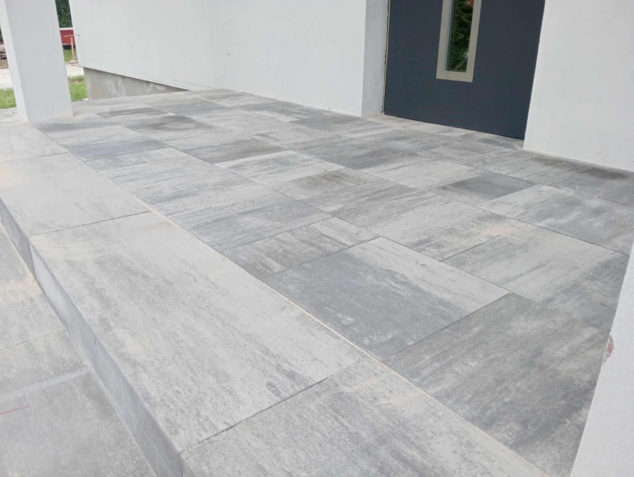 Wykończone schody wejściowe do domu z szarych, prostokątnych płyt betonowych o delikatnej fakturze, prowadzące do ciemnoszarych drzwi z małym okienkiem.