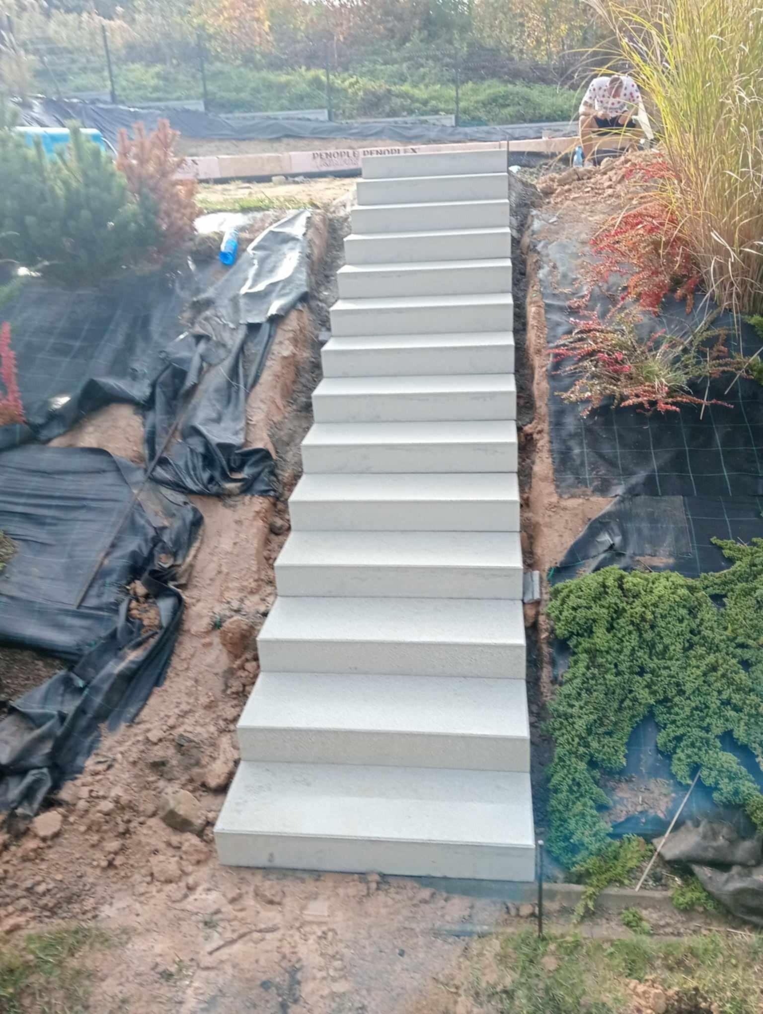 Nowe, jasnoszare schody zewnętrzne z betonu prowadzące w górę po zboczu; widoczne prace ziemne, czarna agrowłóknina i roślinność ozdobna.