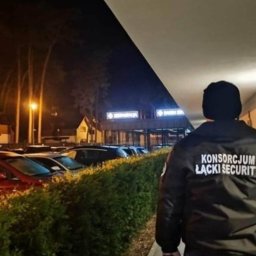 Grupy Łącki sp. zoo. - Ochroniarz w czarnej kurtce z napisem 'KONSORCJUM ŁĄCKI SECURITY' na plecach, patrolujący parking przed budynkiem oświetlonym światłem nocnym.