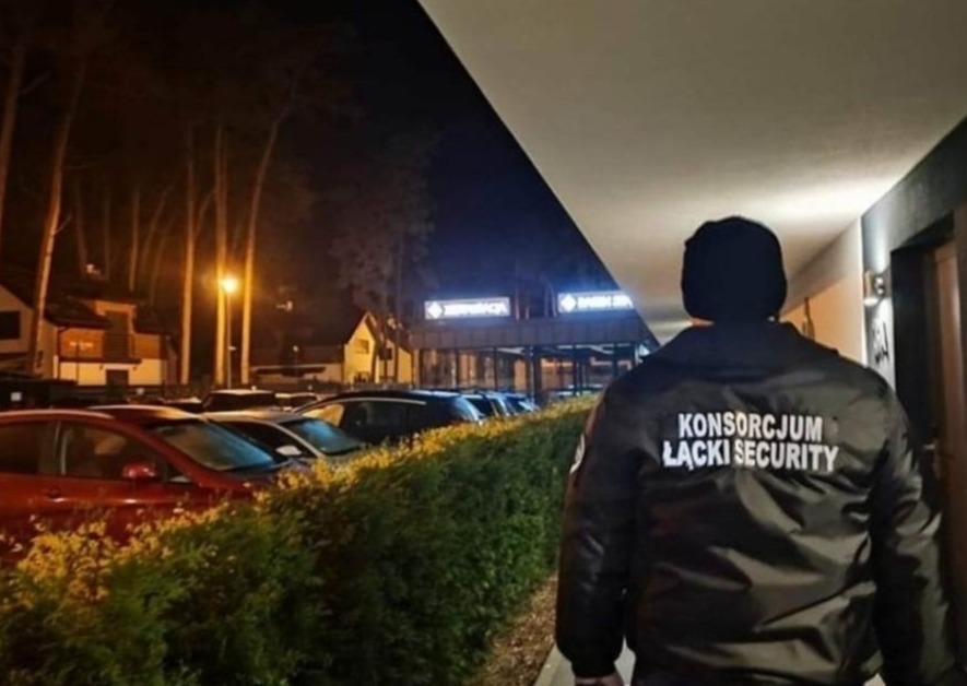 Ochroniarz w czarnej kurtce z napisem 'KONSORCJUM ŁĄCKI SECURITY' na plecach, patrolujący parking przed budynkiem oświetlonym światłem nocnym.