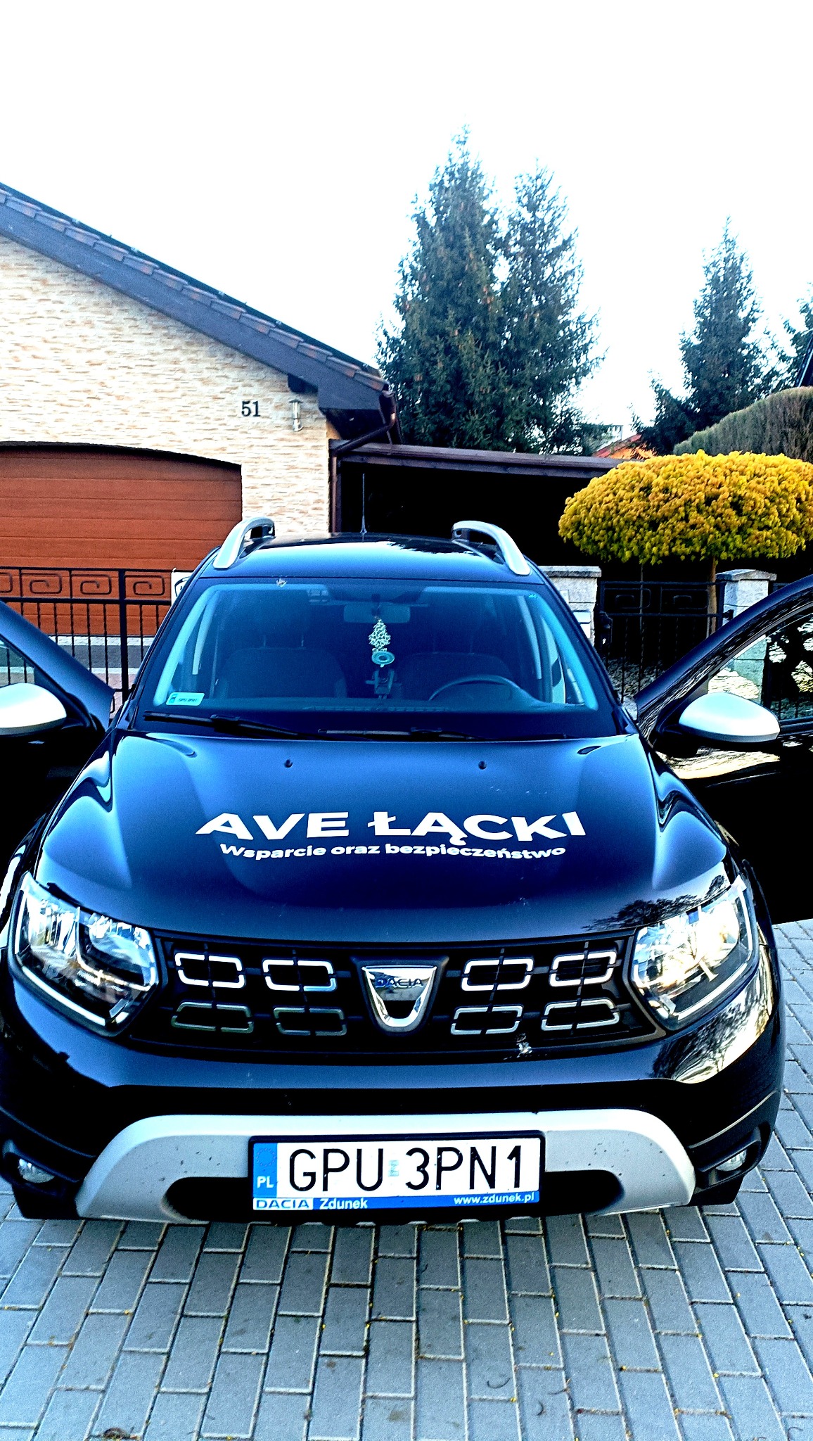 Czarny samochód marki Dacia z otwartymi drzwiami, z napisem na masce 'AVE ŁĄCKI Wsarcie oraz bezpieczeństwo', zaparkowany na brukowanej nawierzchni przed domem.