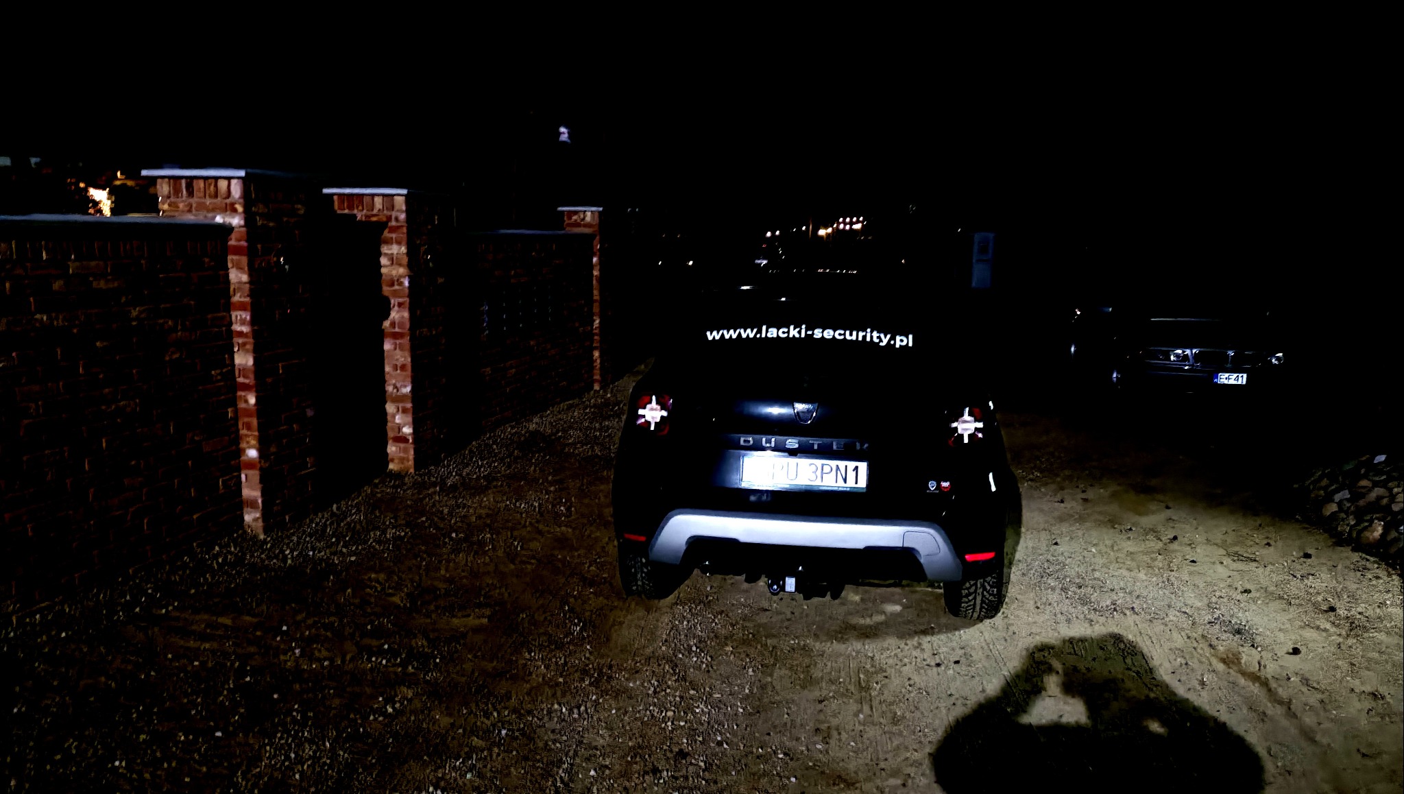Czarny samochód Dacia Duster z napisem 'www.lacki-security.pl' na klapie bagażnika, zaparkowany nocą na żwirowej drodze obok ceglanego ogrodzenia, oświetlony reflektorami. Widoczny cień osoby...