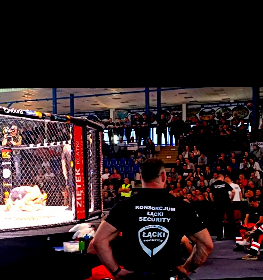 Ochroniarz z logiem 'Łącki Security' na czarnej koszulce, stojący tyłem do kamery podczas wydarzenia sportowego MMA, z widoczną klatką i publicznością w tle.