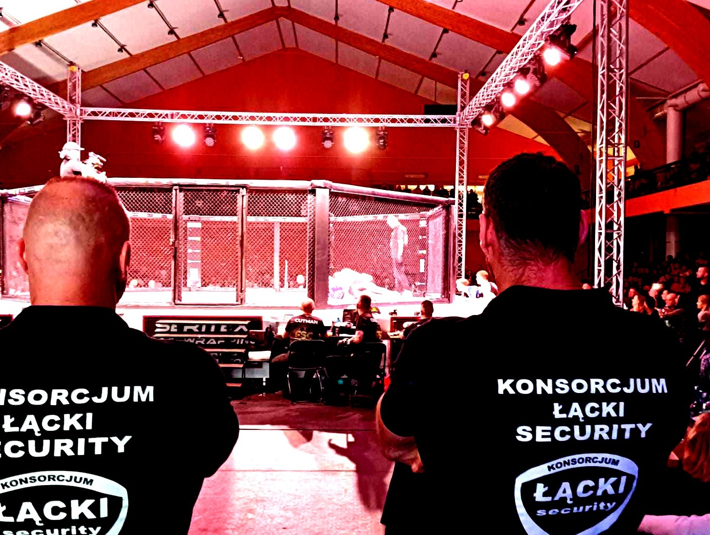 Dwóch ochroniarzy w czarnych koszulkach z napisem 'Konsorcjum Łącki Security' stoi tyłem do widza, obserwując klatkę MMA podczas wydarzenia sportowego. W tle widoczni widzowie i oświetlenie sceniczne.