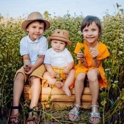 Zagroda Pod Górą - Trójka uśmiechniętych dzieci w kapeluszach siedzi na drewnianej skrzyni na tle kwitnącego pola gryki, starsze dzieci w sandałach, najmłodsze dziecko w żółtych spodenkach