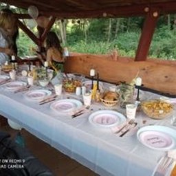 Zagroda Pod Górą - Uroczysty stół nakryty białym obrusem, z różowymi talerzami, srebrnymi sztućcami i dekoracjami kwiatowymi pod drewnianą altaną. Widoczne dwie osoby przygotowujące się do wydarzenia.
