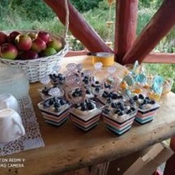 Zagroda Pod Górą - Stół z drewnianym blatem, na którym znajdują się desery w kwadratowych naczyniach z czarno-biało-czerwonymi paskami, udekorowane borówkami, obok białe filiżanki, koszyk z jabłkami i limonkami...
