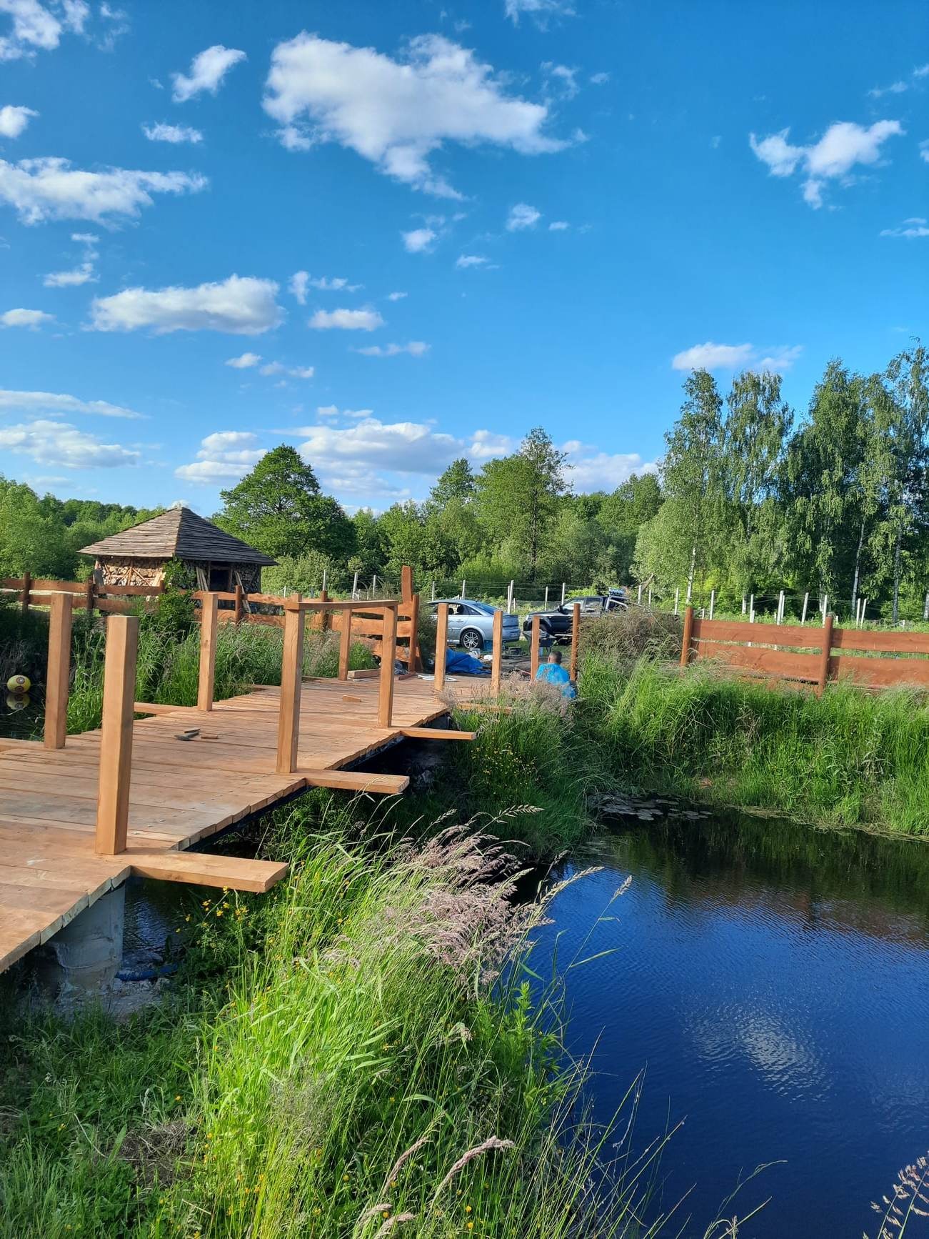 Drewniany mostek w trakcie budowy nad ciemną rzeką, w tle altana z drewnianym dachem i zaparkowane samochody, słoneczny dzień.