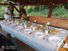 Uroczysty stół nakryty białym obrusem, z różowymi talerzami, srebrnymi sztućcami i dekoracjami kwiatowymi pod drewnianą altaną. Widoczne dwie osoby przygotowujące się do wydarzenia.