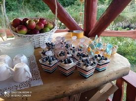 Stół z drewnianym blatem, na którym znajdują się desery w kwadratowych naczyniach z czarno-biało-czerwonymi paskami, udekorowane borówkami, obok białe filiżanki, koszyk z jabłkami i limonkami...