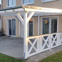 Biała drewniana pergola z balustradą w kształcie krzyży, przylegająca do domu z beżową elewacją, widoczne okna i drzwi balkonowe, na pierwszym planie trawnik.