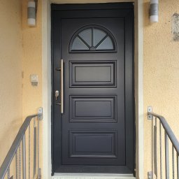 Holzmet s.c. S.Maślany, M. Maślany - Czarna, elegancka drzwi wejściowe z półokrągłym przeszkleniem u góry, złoty uchwyt i klucz w zamku, widoczne fragmenty metalowej balustrady i lampy ścienne na fasadzie o nierównej strukturze.