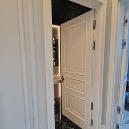 Holzmet s.c. S.Maślany, M. Maślany - Otwarta biała, płycinowa konstrukcja drzwi wewnętrznych z widocznymi zawiasami, prowadząca do łazienki z czarnymi marmurowymi płytkami i podświetlanym lustrem.