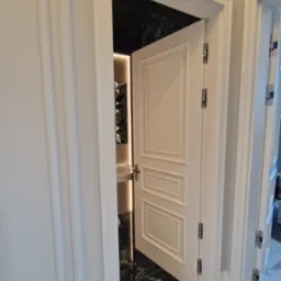 Otwarta biała, płycinowa konstrukcja drzwi wewnętrznych z widocznymi zawiasami, prowadząca do łazienki z czarnymi marmurowymi płytkami i podświetlanym lustrem.