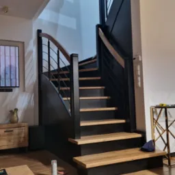 Nowoczesne schody z czarnymi bokami i drewnianymi stopniami oraz metalową balustradą w jasnym wnętrzu. Widoczny fragment podłogi z desek i puszka farby w sprayu.