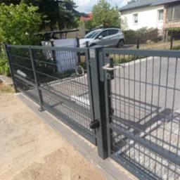 Nowoczesna, szara, metalowa brama panelowa z widocznym zamkiem i klamką, prowadząca na podjazd wyłożony kostką brukową, w tle dom i samochód.