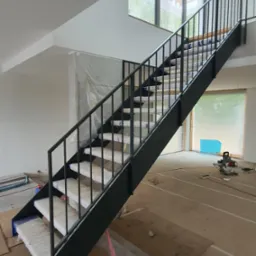 Nowoczesne schody z czarną metalową balustradą i schodami zabezpieczonymi folią, widoczne podczas montażu w jasnym wnętrzu z dużym oknem.