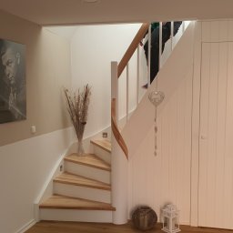 Holzmet s.c. S.Maślany, M. Maślany - Białe schody z drewnianymi stopniami i poręczą, ozdobione wazonem z suszonymi kwiatami i dekoracją w kształcie serca, widoczne fragmenty osoby wchodzącej po schodach.