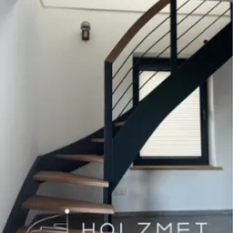 Spiralne schody z drewnianymi stopniami i czarną metalową konstrukcją, widoczne od dołu, z oknem w tle i lampą na ścianie. Na pierwszym planie logo firmy.