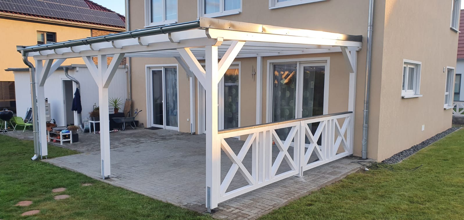 Biała drewniana pergola z balustradą w kształcie krzyży, przylegająca do domu z beżową elewacją, widoczne okna i drzwi balkonowe, na pierwszym planie trawnik.