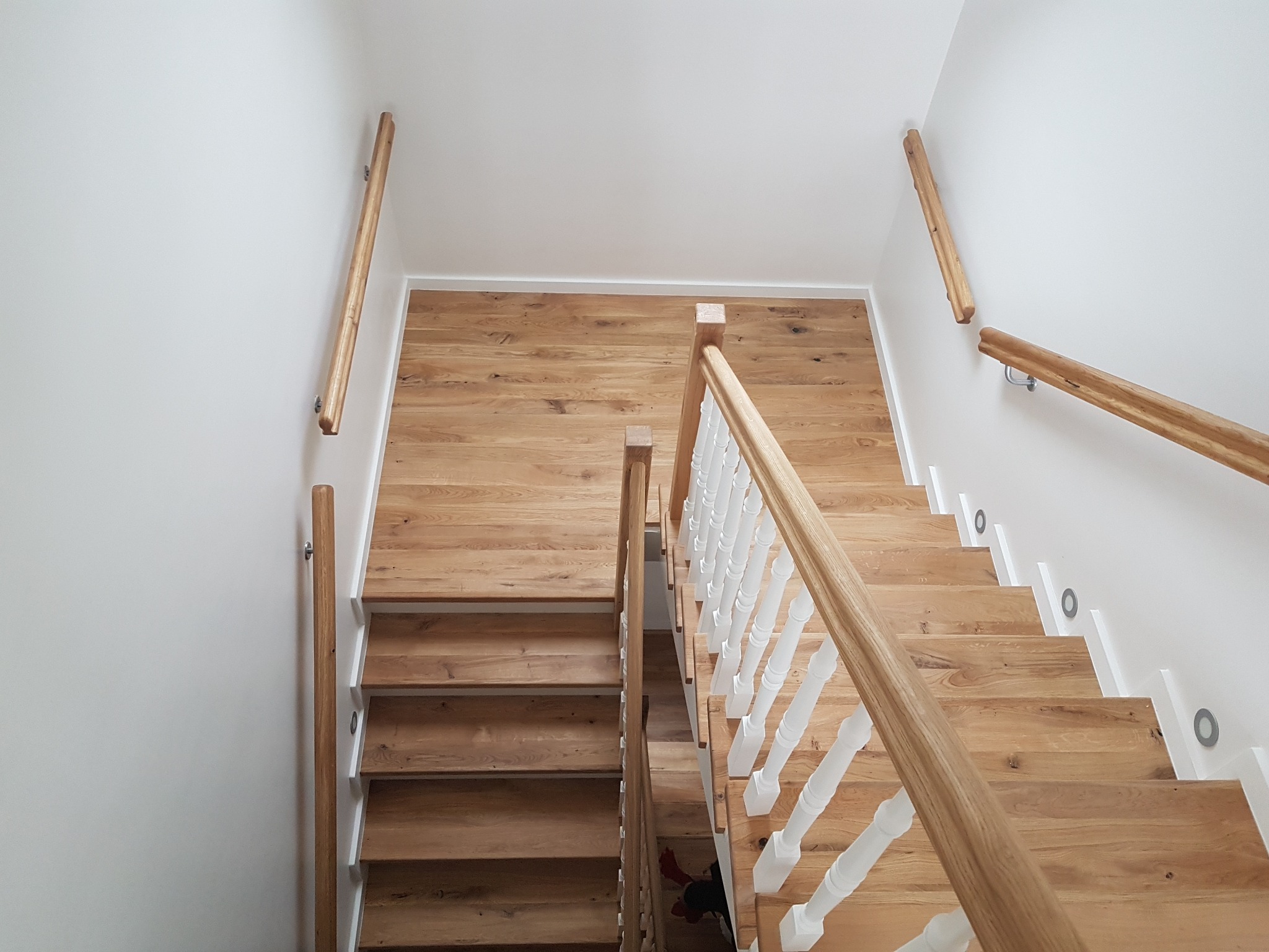 Widok z góry na drewniane schody z białymi balustradami i drewnianą poręczą, zintegrowane oświetlenie w ścianie, oraz drewniane wykończenie ściany na podeście.