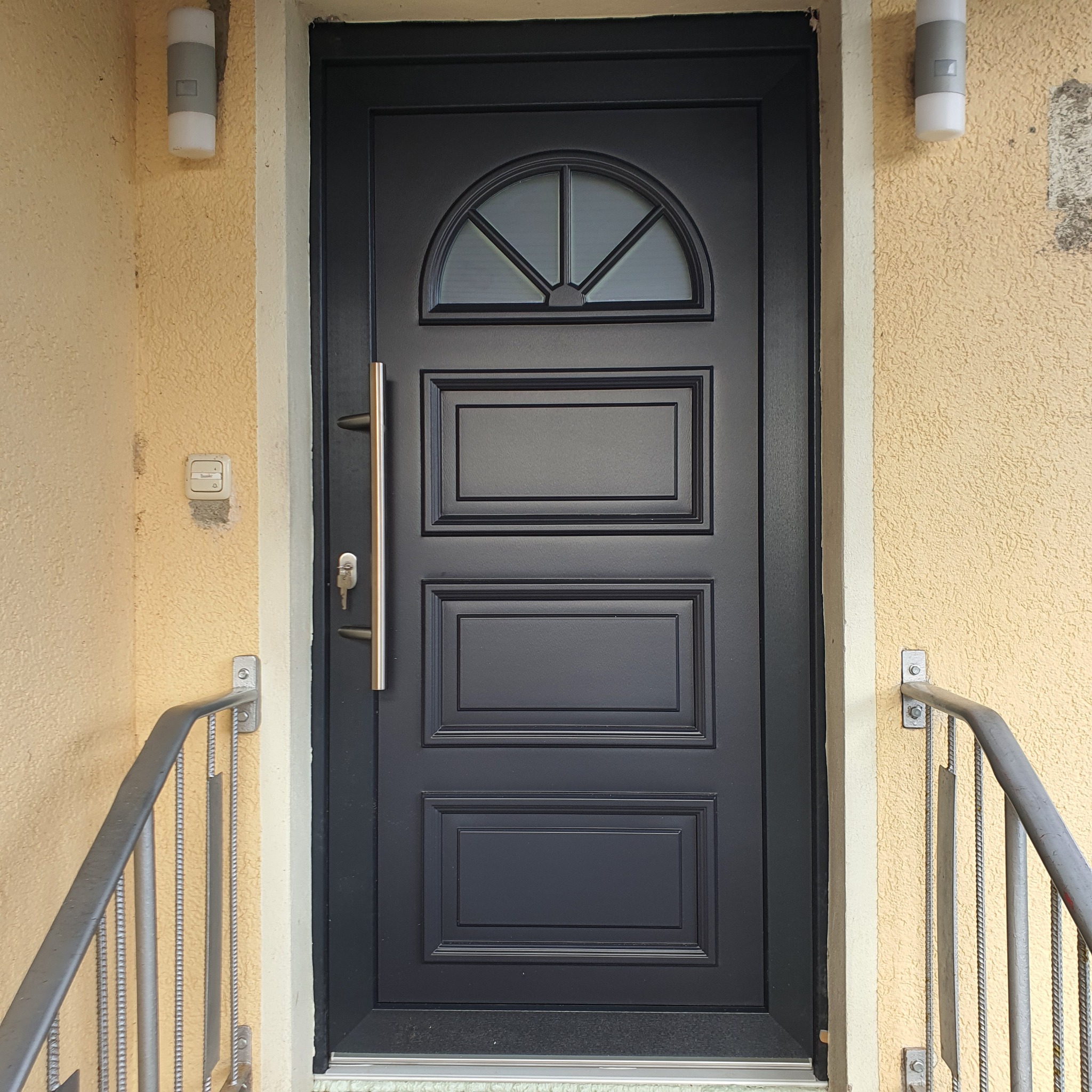 Czarna, elegancka drzwi wejściowe z półokrągłym przeszkleniem u góry, złoty uchwyt i klucz w zamku, widoczne fragmenty metalowej balustrady i lampy ścienne na fasadzie o nierównej strukturze.