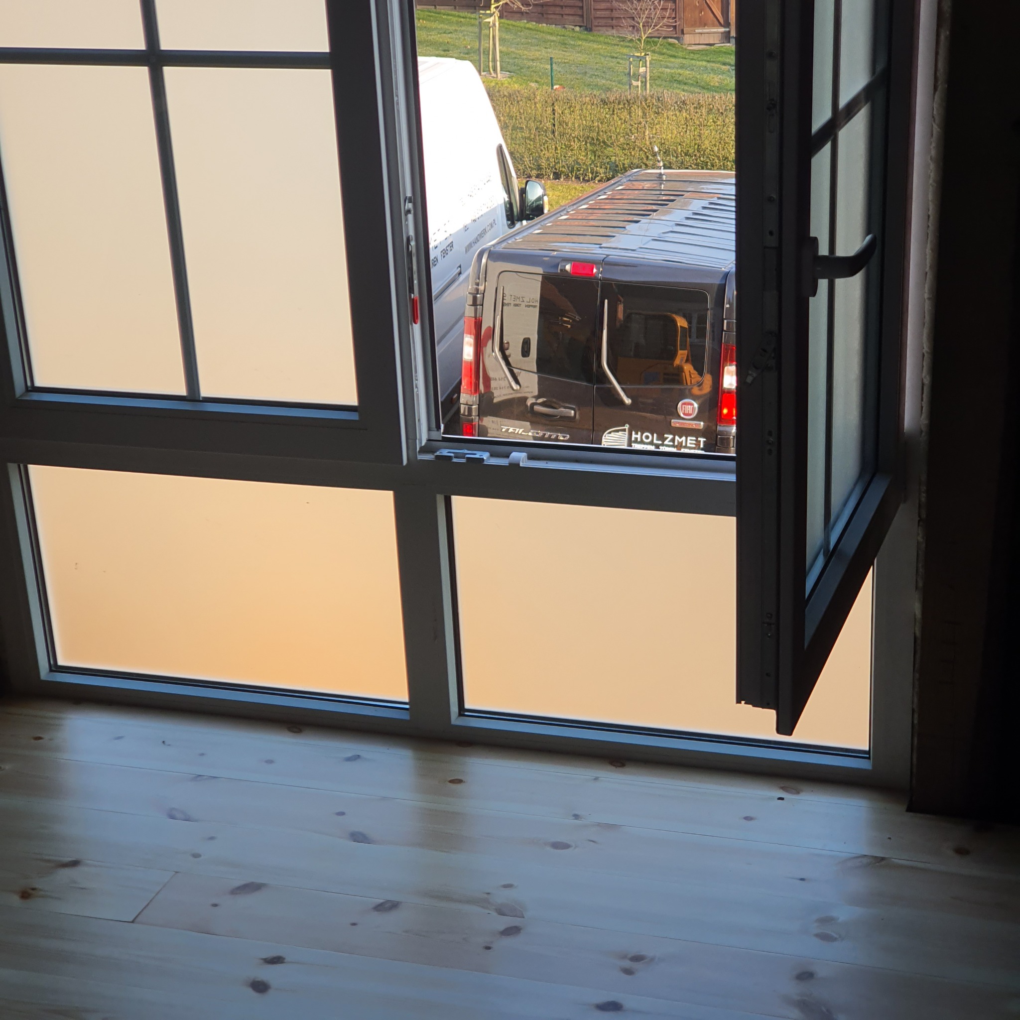 Otwarta okno z szybami o mlecznym wykończeniu, przez które widać zaparkowane samochody dostawcze na zewnątrz, drewniana podłoga wewnątrz pomieszczenia.