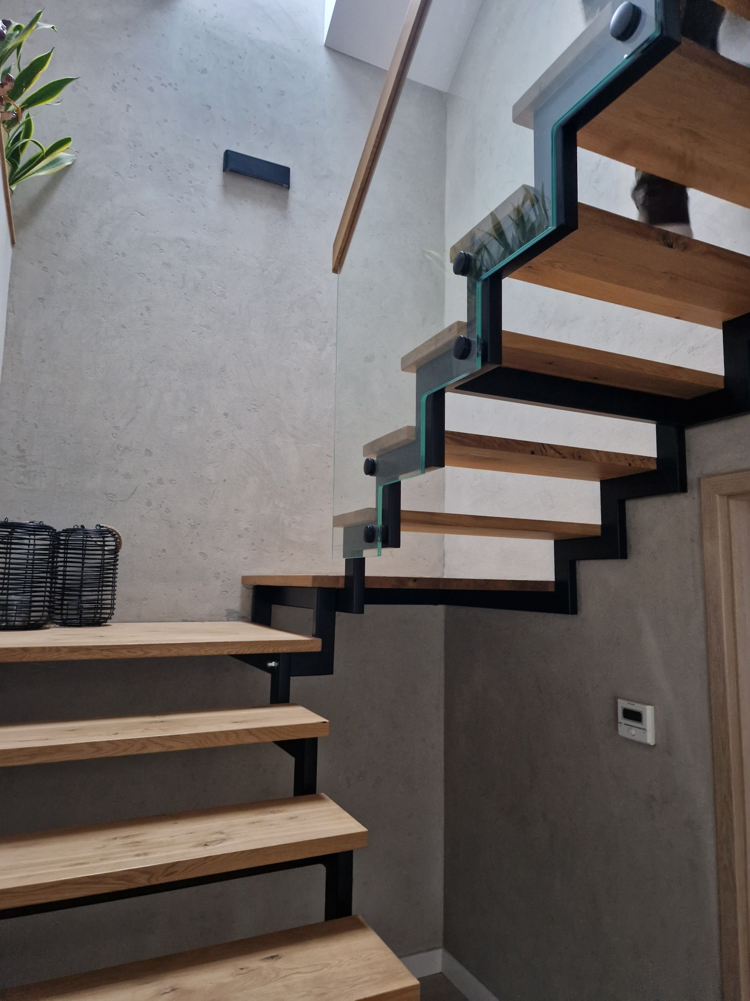 Nowoczesne schody z drewnianymi stopniami, czarną metalową konstrukcją i szklaną balustradą, oświetlone światłem wpadającym z góry, z dekoracyjnymi koszykami na dolnym stopniu i minimalistycznym...