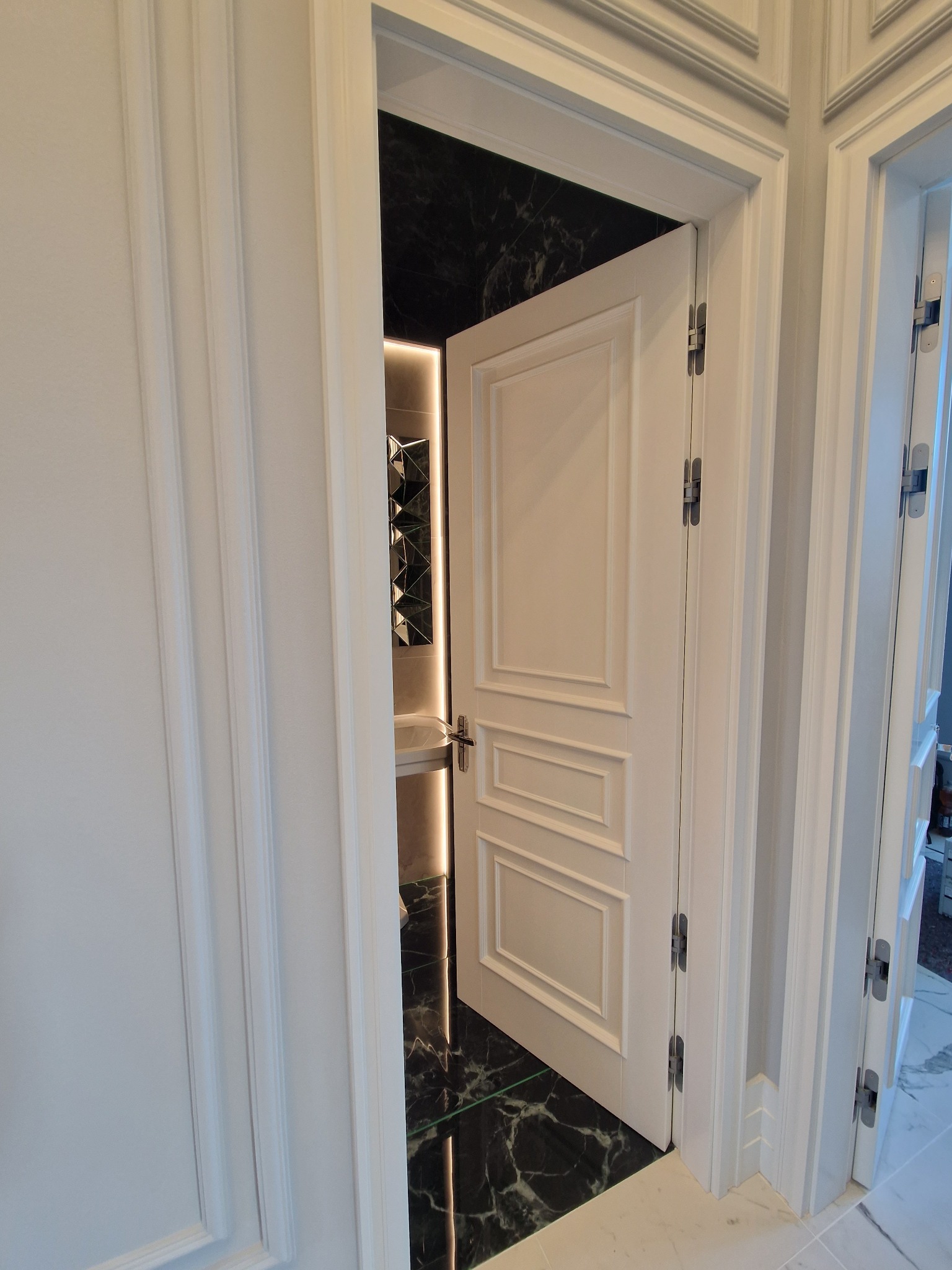 Otwarta biała, płycinowa konstrukcja drzwi wewnętrznych z widocznymi zawiasami, prowadząca do łazienki z czarnymi marmurowymi płytkami i podświetlanym lustrem.
