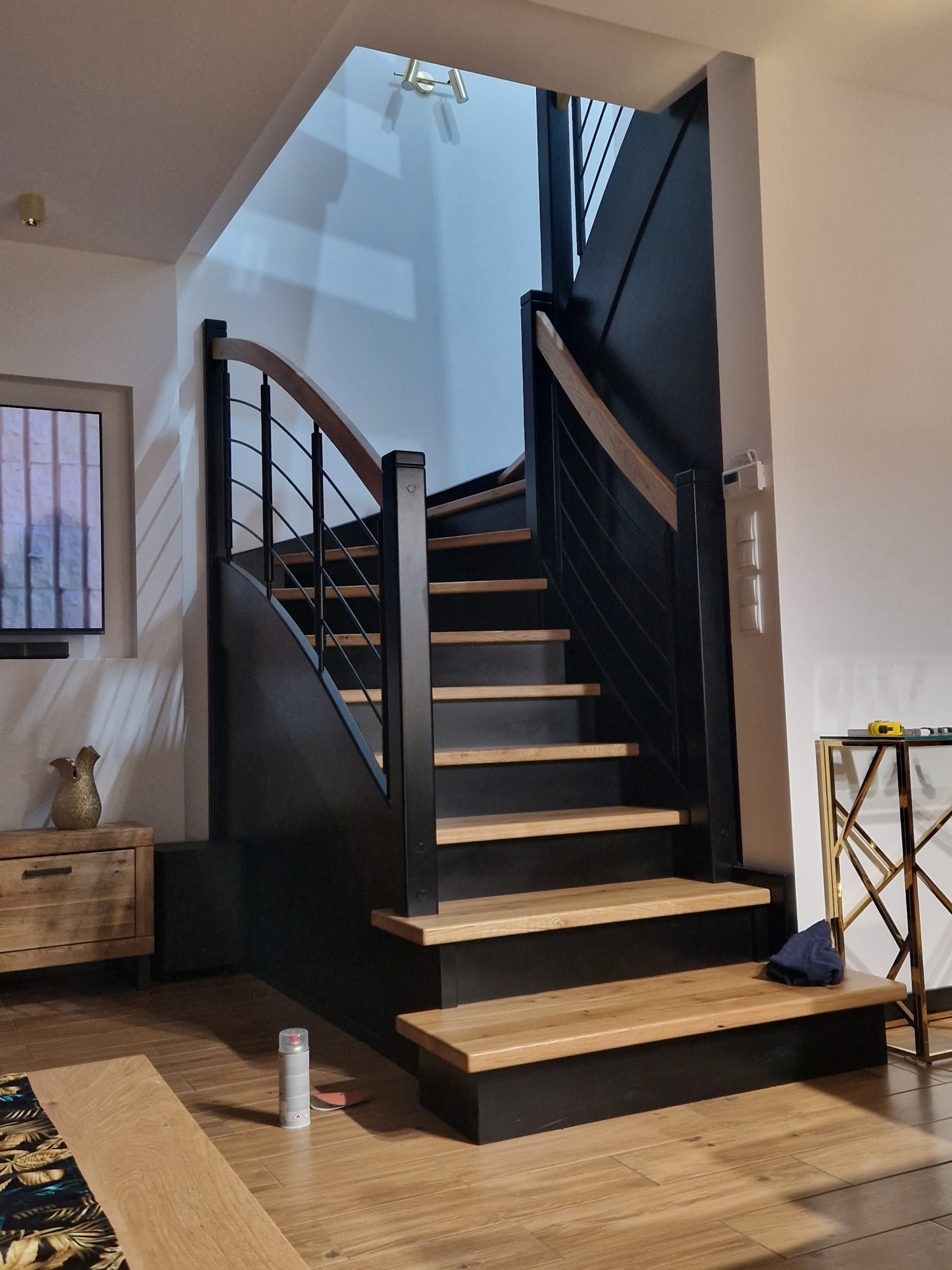 Nowoczesne schody z czarnymi bokami i drewnianymi stopniami oraz metalową balustradą w jasnym wnętrzu. Widoczny fragment podłogi z desek i puszka farby w sprayu.