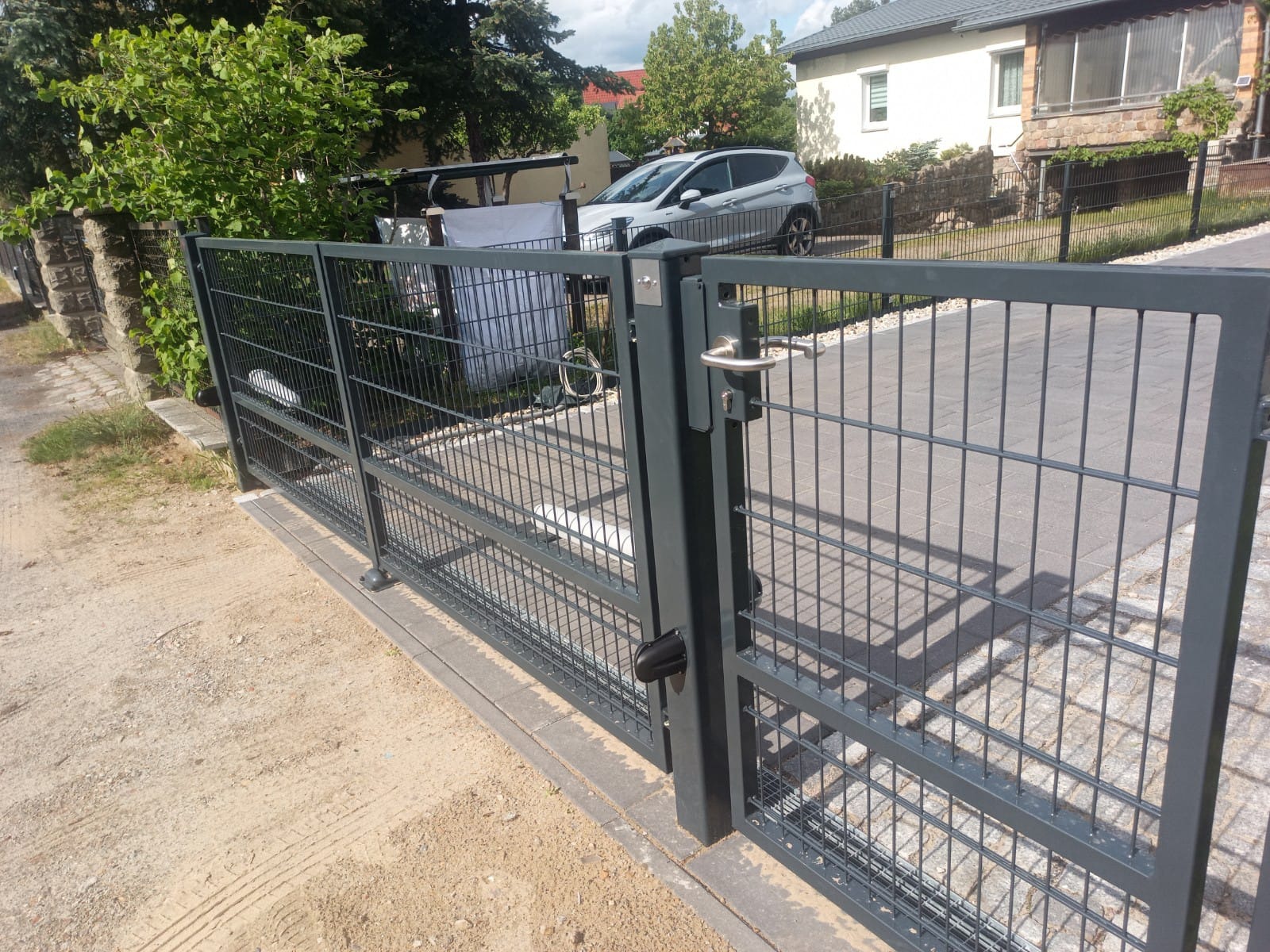 Nowoczesna, szara, metalowa brama panelowa z widocznym zamkiem i klamką, prowadząca na podjazd wyłożony kostką brukową, w tle dom i samochód.